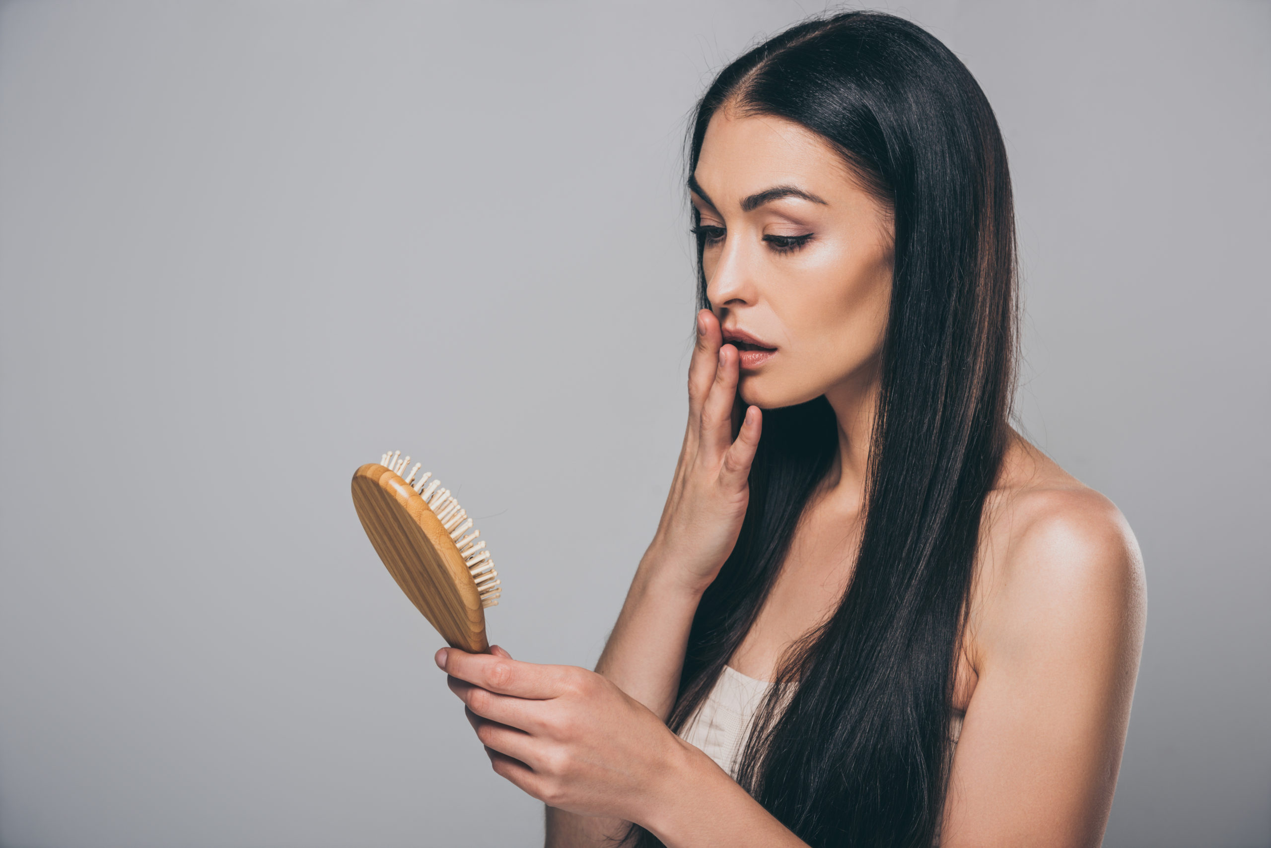 shocked young brunette woman holding hairbrush isolated on grey, hair loss concept
